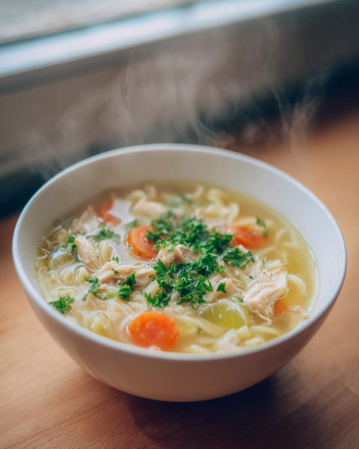 A steaming white bowl of amazing penicillin soup featuring shredded chicken, egg noodles, carrots, and fresh parsley garnish.