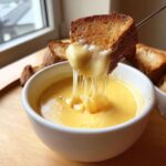 A piece of toasted bread is dipped into a white bowl of hot, melted cheese fondue recipe, showing long cheese pulls.