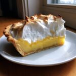 A thick slice of lemon meringue pie showing the bright yellow filling and tall, toasted meringue topping.