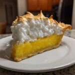 A close-up of a perfect slice of lemon meringue pie showing bright yellow filling and toasted meringue peaks.