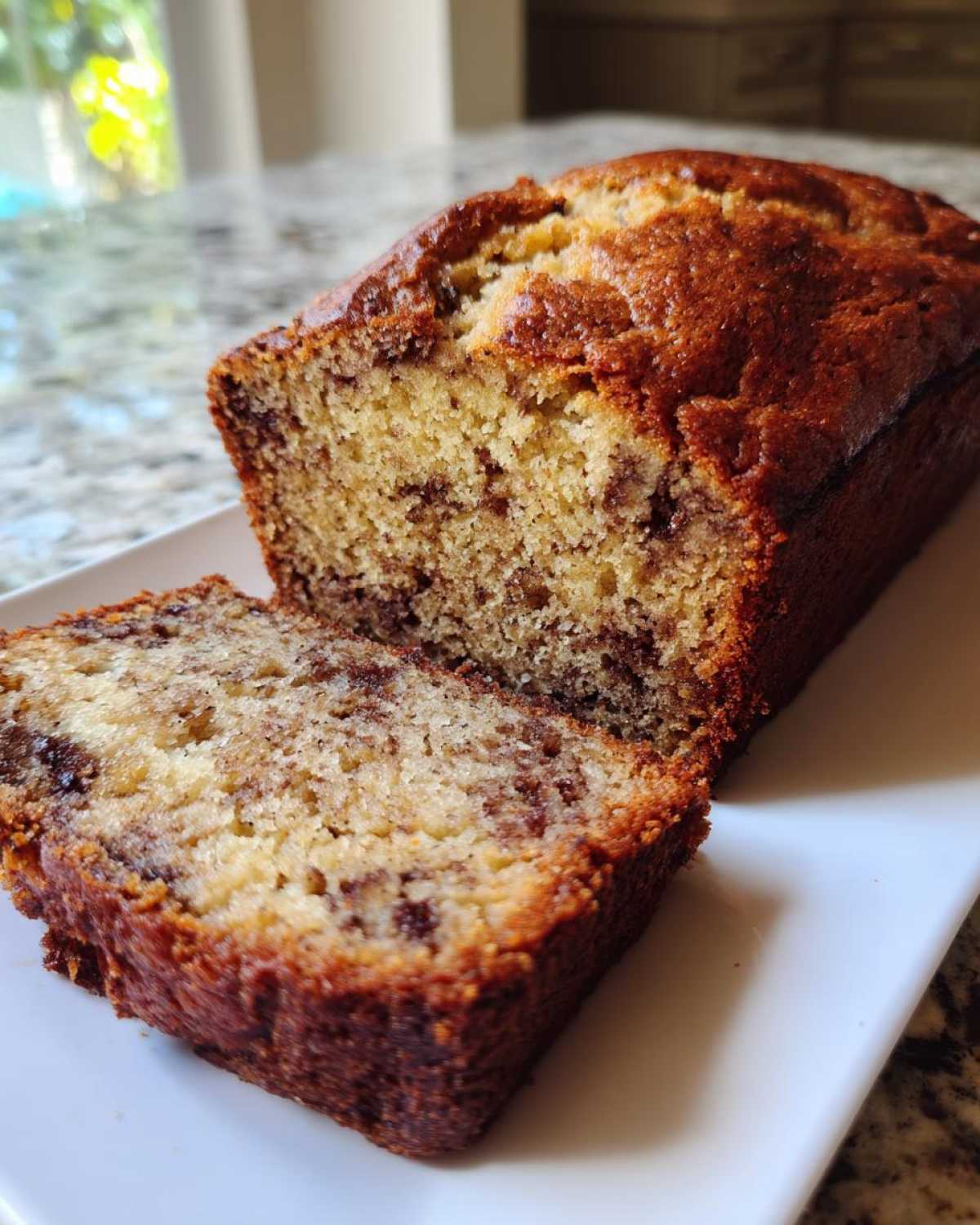 A loaf of moist sugar free banana bread with one thick slice cut and resting against the main loaf on a white platter.