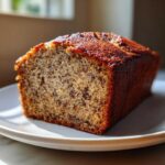 A close-up, golden-brown slice of moist sugar free banana bread sitting on a white plate, showing speckles of banana.