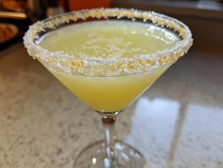 Close-up of a bright yellow lemon drop cocktail in a martini glass with a thick sugar and lemon zest rim.