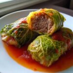 Three homemade cabbage rolls served on a white plate, one cut open to show the savory meat filling inside, covered in tomato sauce.