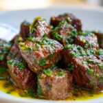 Close-up of tender crockpot steak bites covered in a bright green herb and garlic sauce, resting in yellow juices.