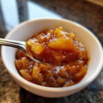 A spoonful of thick, spiced apple pie filling lifted from a white bowl, showing chunky apple pieces.