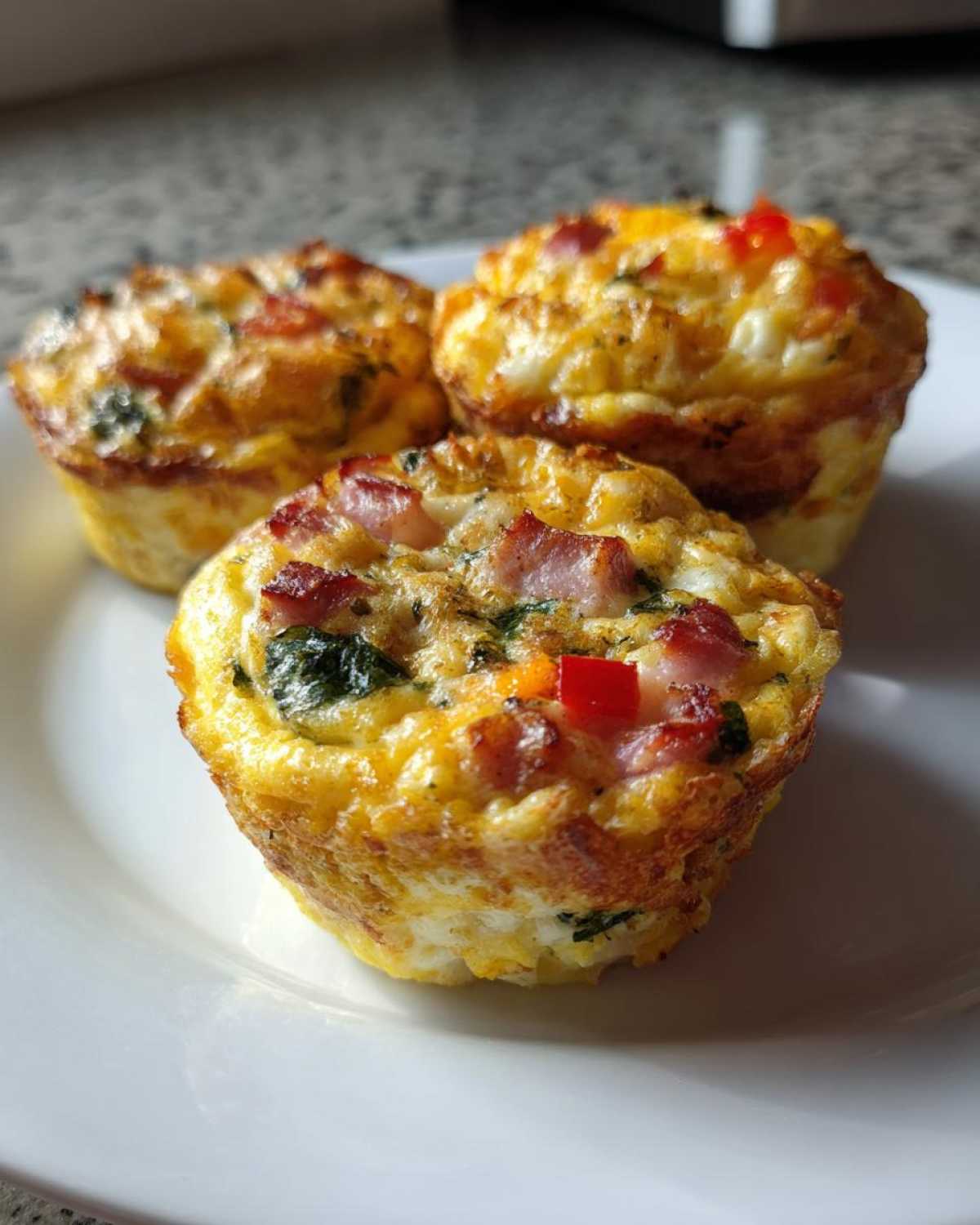 Three perfectly baked Egg Muffin Cups topped with bacon, spinach, and diced red pepper on a white plate.