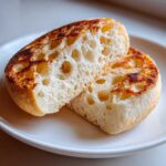 A toasted english muffin, split in half, showing its signature open, airy crumb structure on a white plate.