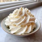 A close-up of fluffy, pale yellow vanilla buttercream frosting piped high in a small bowl.