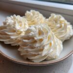 Four swirls of creamy, pale yellow vanilla buttercream frosting piped onto a small ceramic plate.