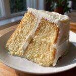 Close-up of a moist slice of vanilla cake recipe layered with thick white buttercream frosting on a white plate.