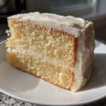 A close-up slice of moist vanilla cake recipe layered with white frosting on a white plate.