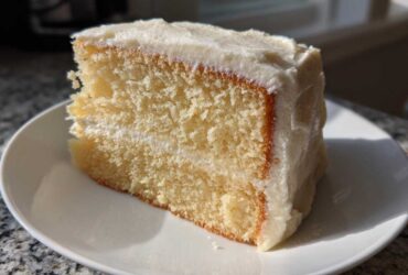 A close-up slice of moist vanilla cake recipe layered with white frosting on a white plate.