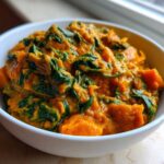 A close-up of a white bowl filled with thick, creamy vegan pumpkin curry featuring chunks of orange squash and wilted green spinach.