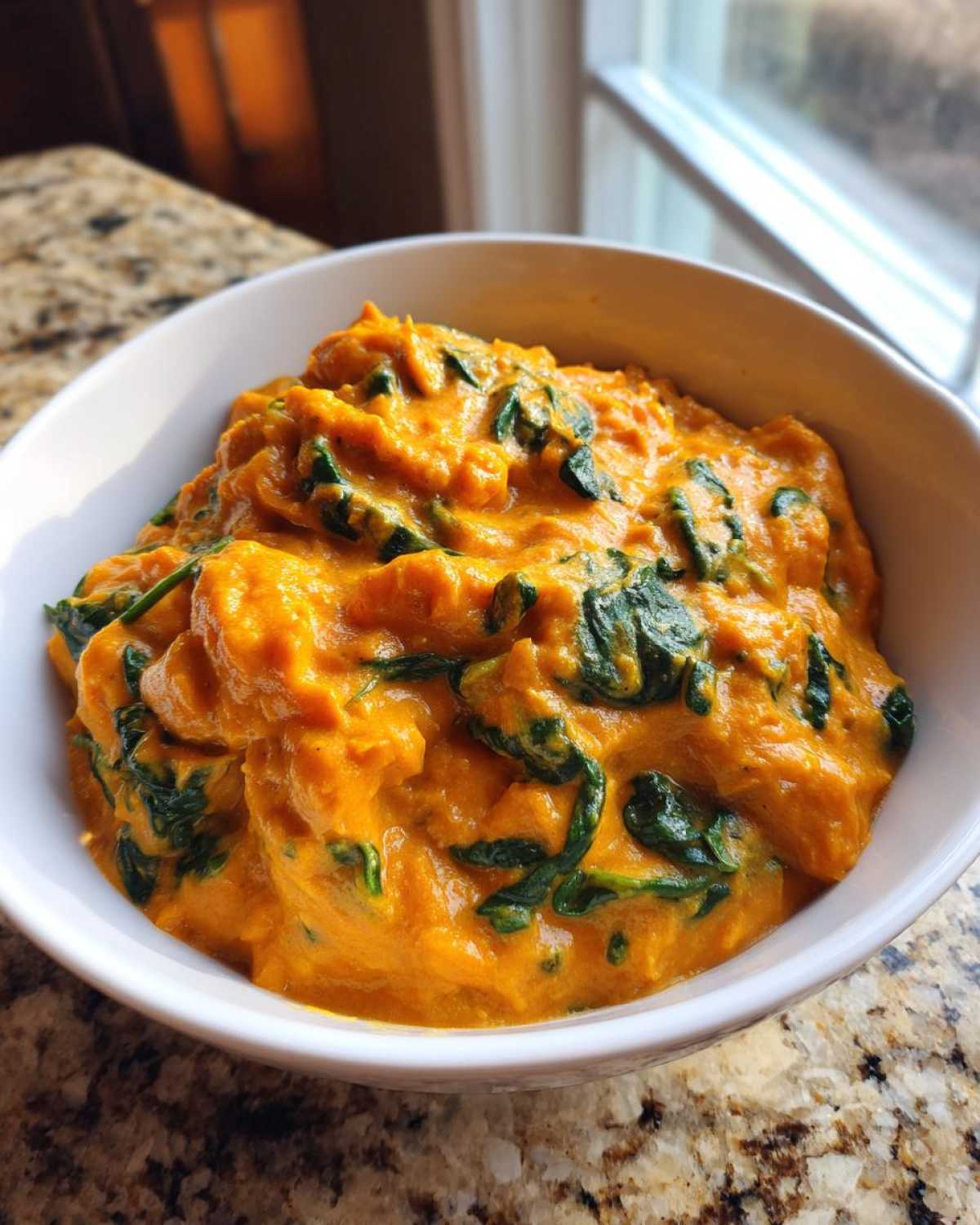 Close-up of a rich, orange vegan pumpkin curry mixed with wilted green spinach served in a white bowl.