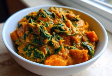 A close-up of a white bowl filled with thick, creamy vegan pumpkin curry featuring chunks of orange squash and wilted green spinach.