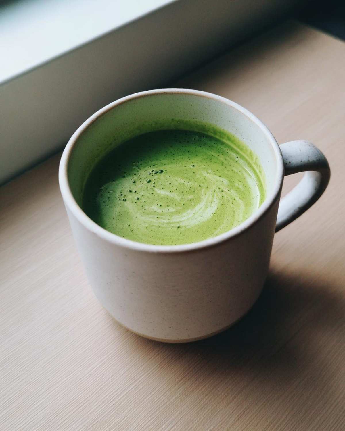 A bright green beverage, likely zucchini soup, with foam swirls in a speckled white mug.