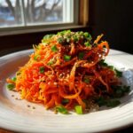 A mound of bright orange raw carrot salad, shredded and topped with sesame seeds and chopped green onions.