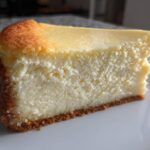 A close-up of a thick, creamy slice of white chocolate cheesecake with a golden-brown top and graham cracker crust.