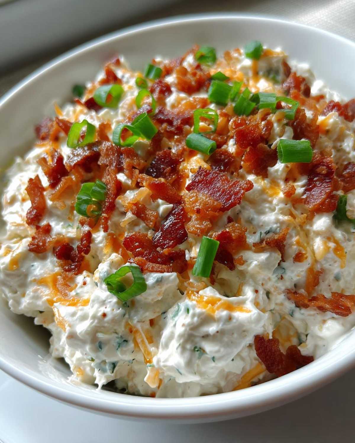 Close-up of a rich cream cheese dip topped generously with crumbled bacon, shredded cheddar, and fresh green onions.