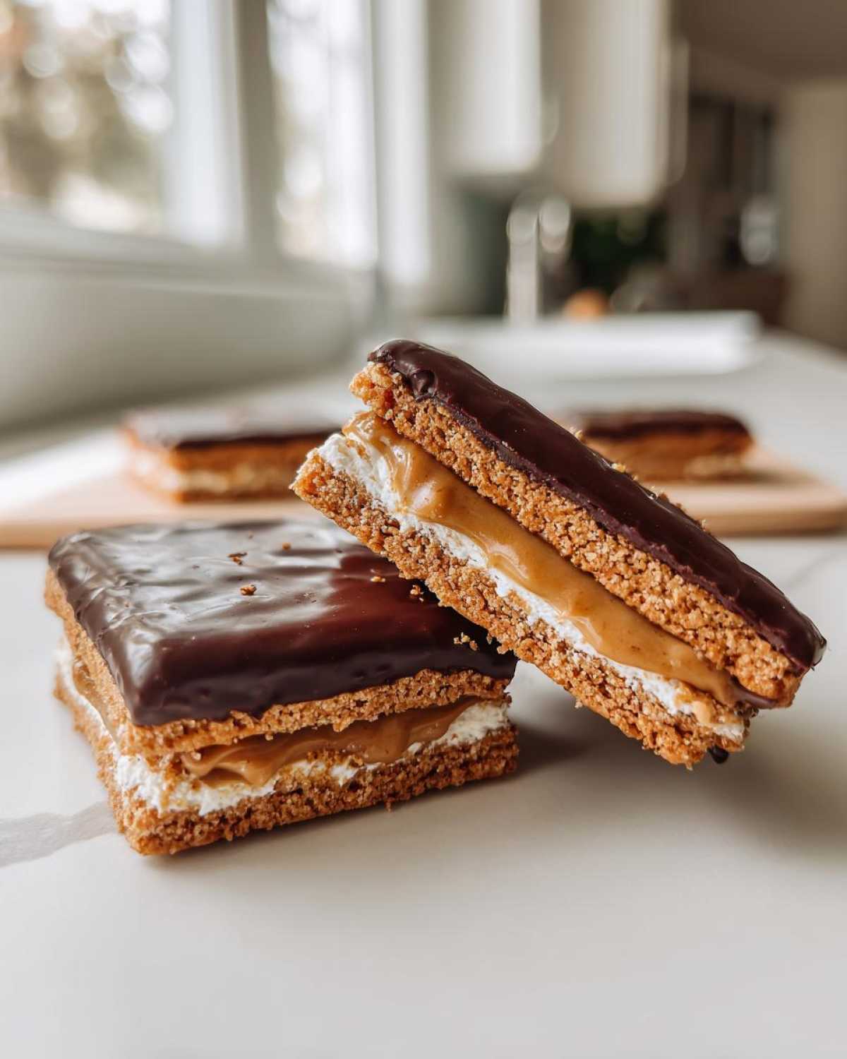 Two layered buckeye graham crackers treats cut in half showing graham cracker base, white filling, peanut butter layer, and chocolate topping.