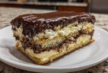 A thick slice of chocolate eclair cake showing layers of pastry, vanilla cream, and rich chocolate frosting.
