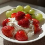 A white bowl filled with cream cheese fruit dip topped with fresh strawberries and green grapes.
