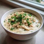 Close-up of a bowl of creamy white chicken chili, topped with shredded chicken, white beans, and fresh parsley.