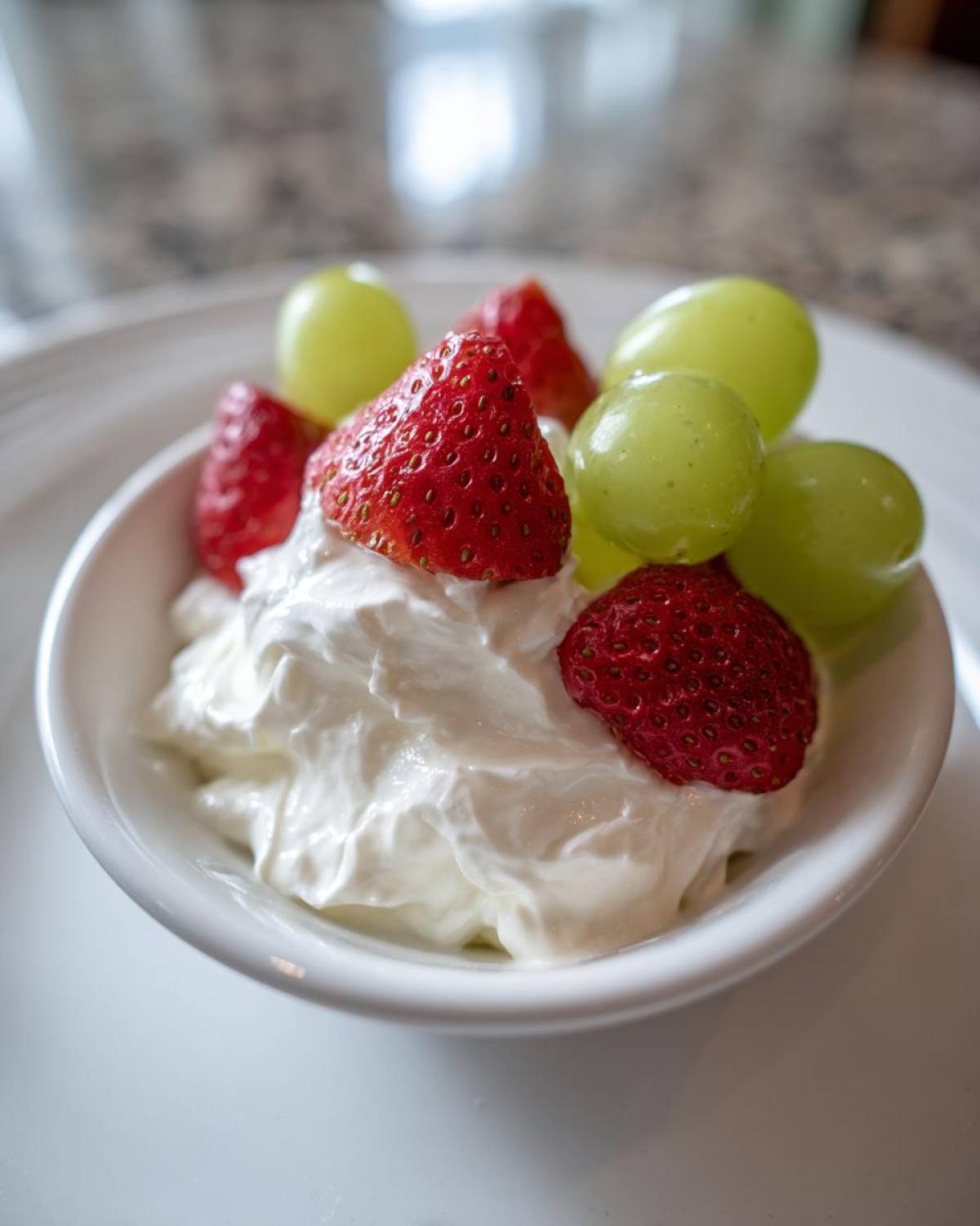 Close-up of a white bowl filled with creamy fruit dip topped with fresh strawberries and green grapes.