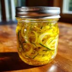 A glass jar filled with bright yellow liquid containing spiralized zucchini and onion slices for bread and butter pickle.