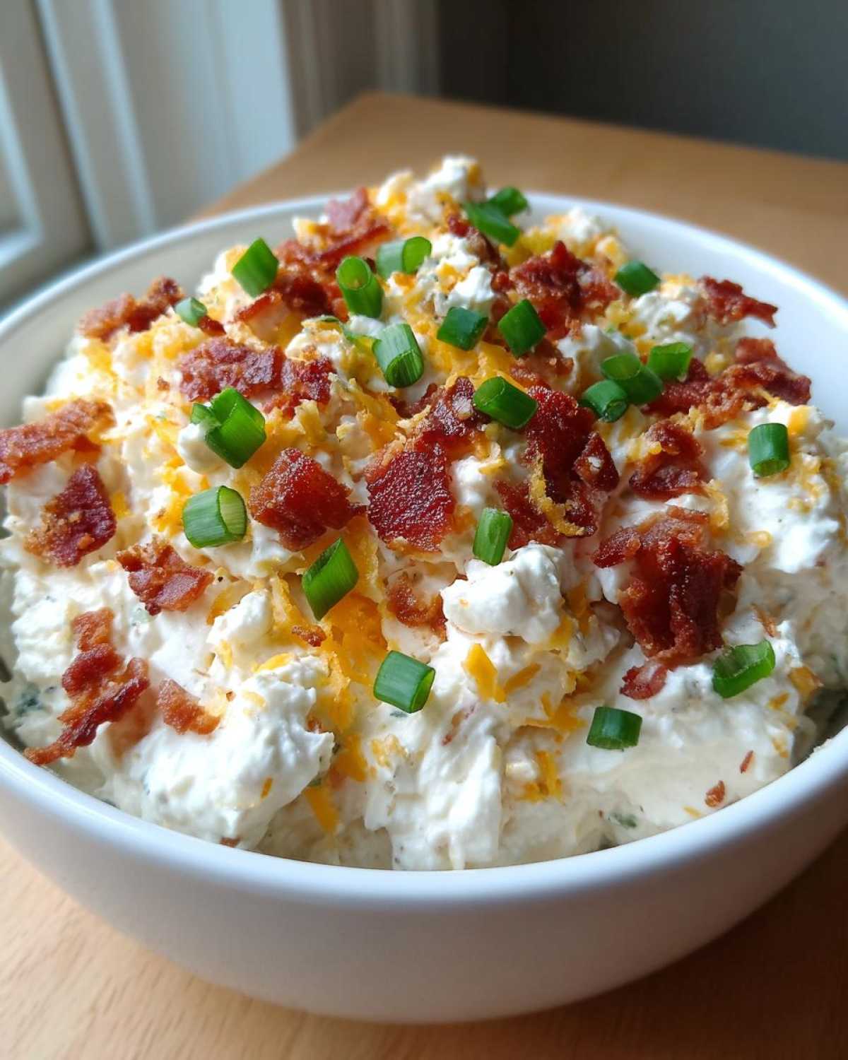 Close-up of a rich cream cheese dip topped generously with bacon bits, shredded cheddar, and fresh green onions.