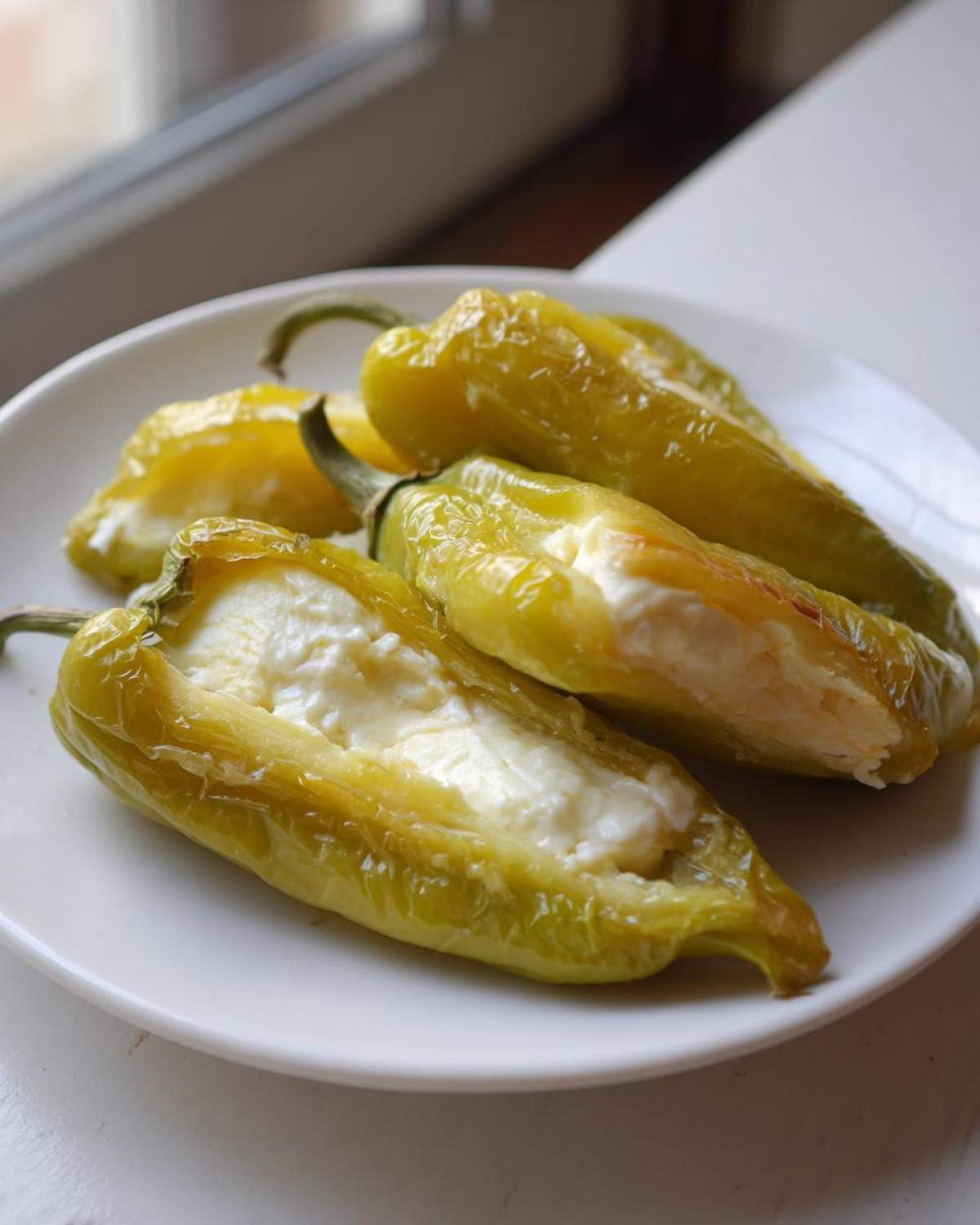 Four baked, light green stuffed banana peppers filled with white cheese mixture, served on a white plate.