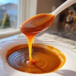 Close-up of thick, glossy date caramel dripping from a white spoon back into a bowl.