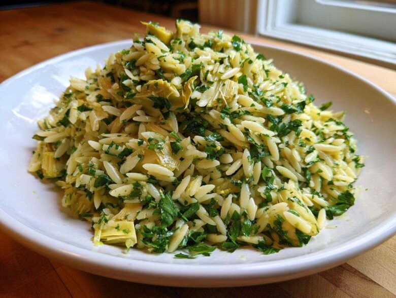 A close-up of a vibrant Artichoke Lemon Orzo Salad mixed with chopped parsley, served in a white bowl.