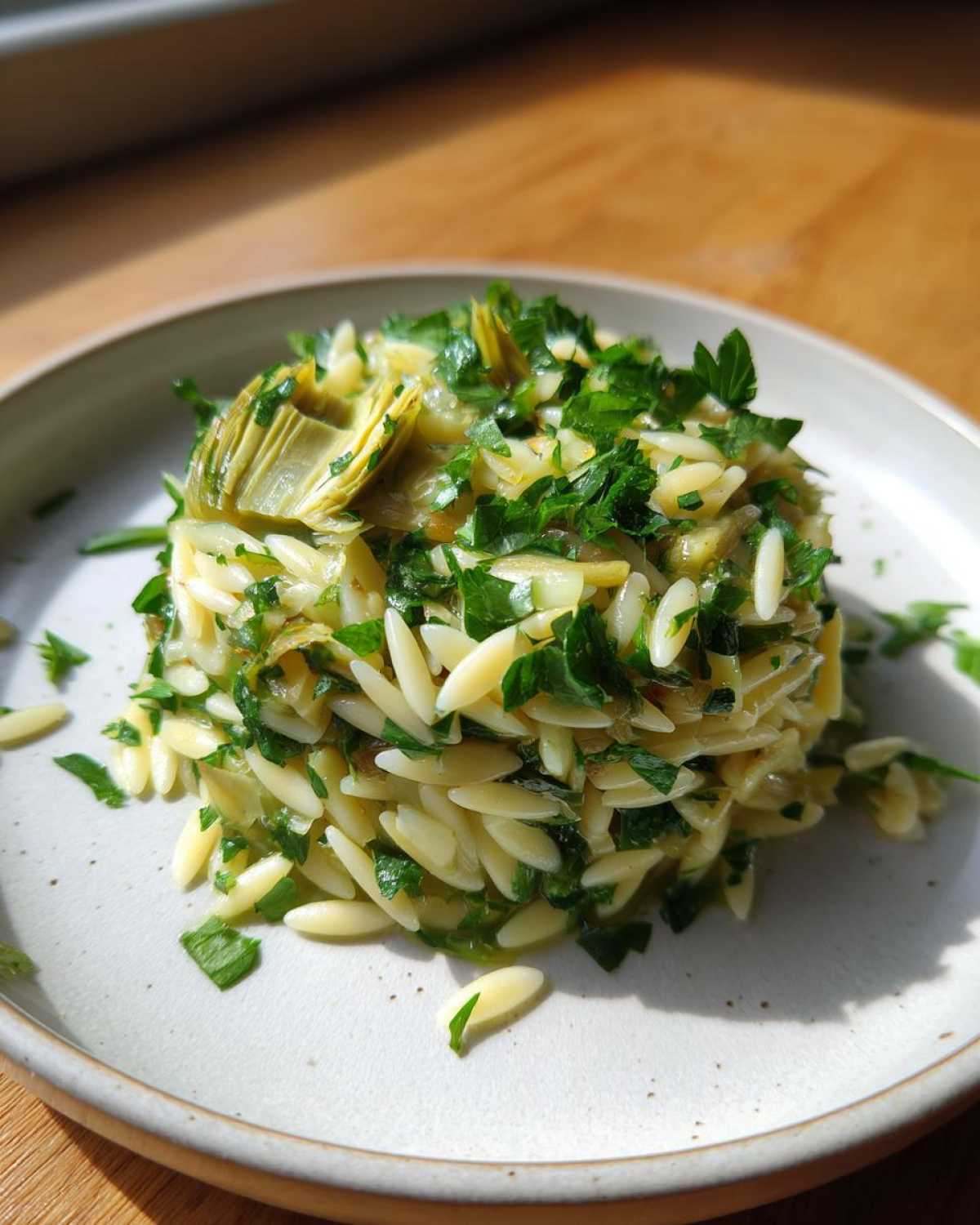 A mound of Artichoke Lemon Orzo Salad mixed with fresh parsley, served on a light-colored plate.