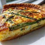 A close-up slice of golden-brown Asparagus Gruyere Quiche with visible asparagus spears on a white plate.