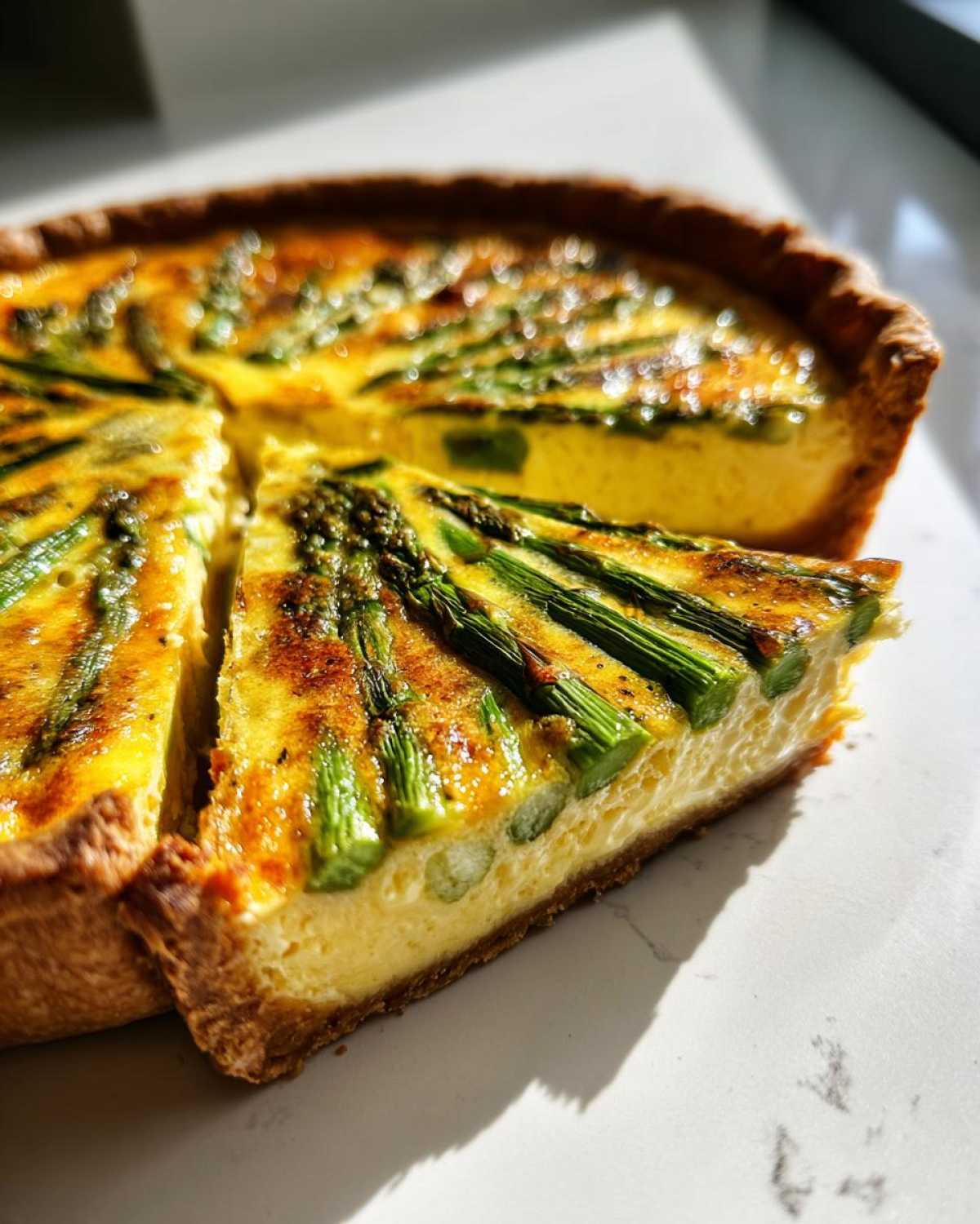 A close-up slice of golden brown Asparagus Gruyere Quiche with visible green asparagus spears on a white plate.