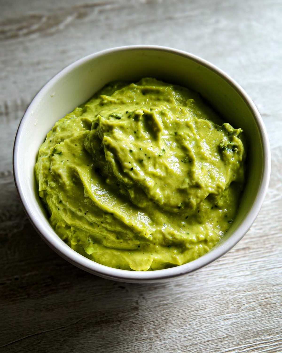 A close-up of thick, bright green Avocado Tomatillo Salsa Verde served in a white bowl on a wooden surface.