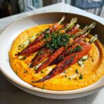 Roasted baby carrots with charred edges served over a vibrant orange harissa yogurt base, garnished with chives.