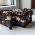 A rich, dark square of Black Sesame Mochi Brownies with a chewy texture and white swirls, served on a white plate.