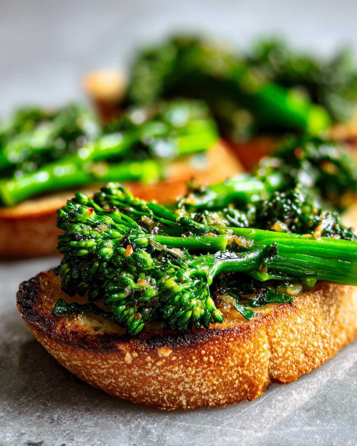 Close-up of a piece of toasted bread topped with bright green, sautéed broccolini for Broccolini Garlic Anchovy Toast.