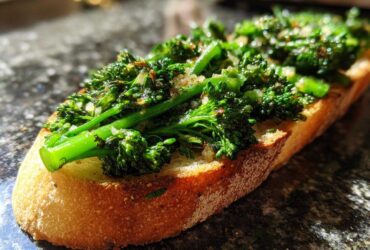 A close-up, sunlit shot of a single slice of Broccolini Garlic Anchovy Toast featuring bright green broccolini on toasted bread.