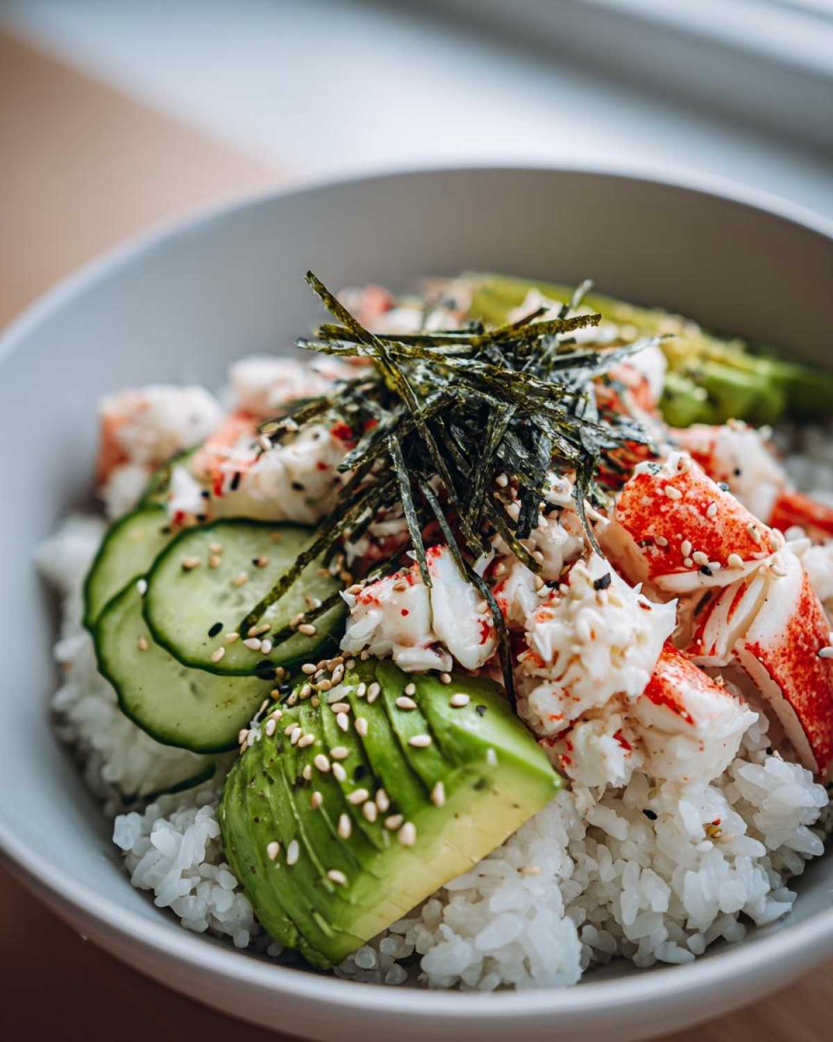 A close-up of a California Sushi Bowl with Crab, featuring white rice, sliced avocado, cucumber, and shredded nori topping.