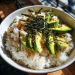 A white bowl filled with white rice, topped with sliced avocado, shredded crab meat, nori strips, and sesame seeds for a California Sushi Bowl with Crab.
