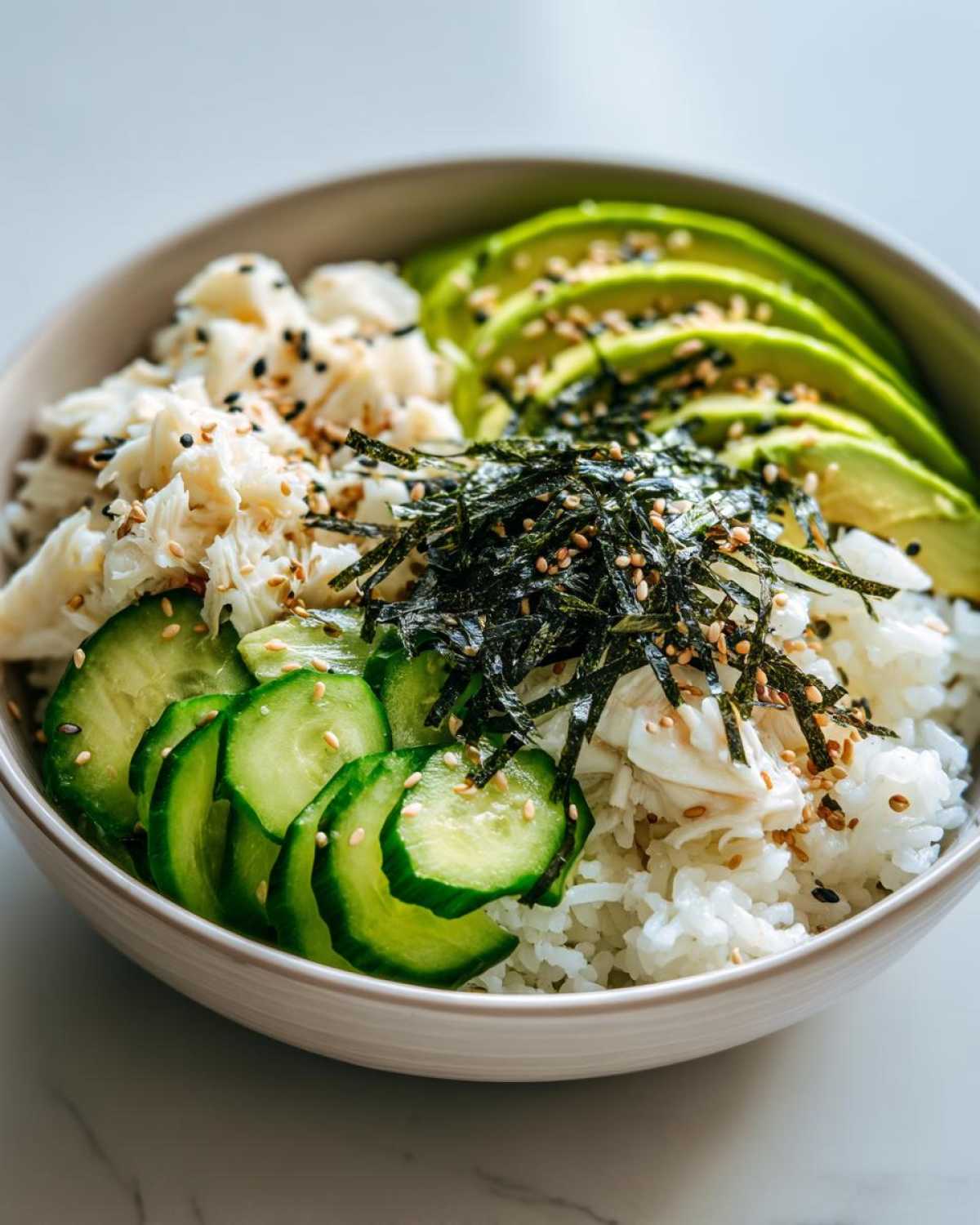 A close-up of a California Sushi Bowl with Crab, featuring rice, sliced avocado, cucumber, shredded nori, and sesame seeds.