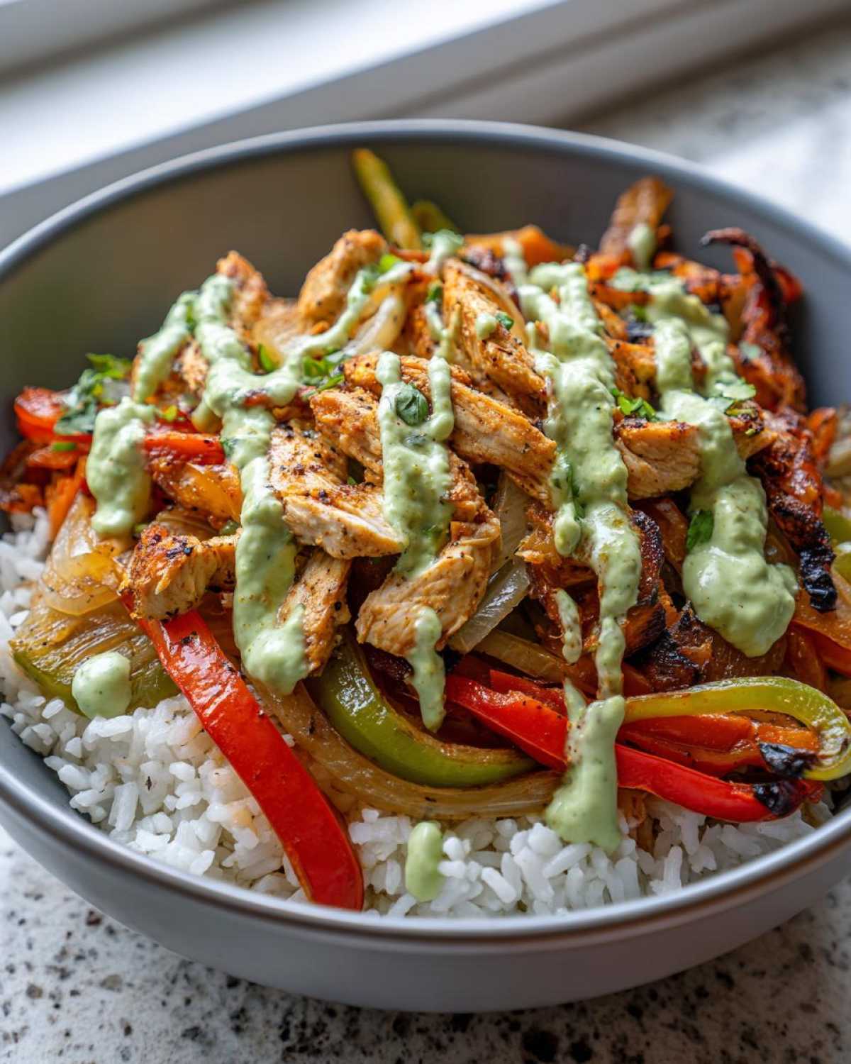 A close-up of a Charred Pepper Fajita Bowl featuring sliced grilled chicken, charred peppers, onions over white rice, drizzled with a light green sauce.