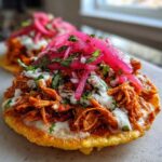 Two crispy Chicken Tinga Tostadas topped with shredded chicken, crema, and bright pink pickled onions.