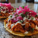 Close-up of two Chicken Tinga Tostadas topped with crema and vibrant pickled onions.