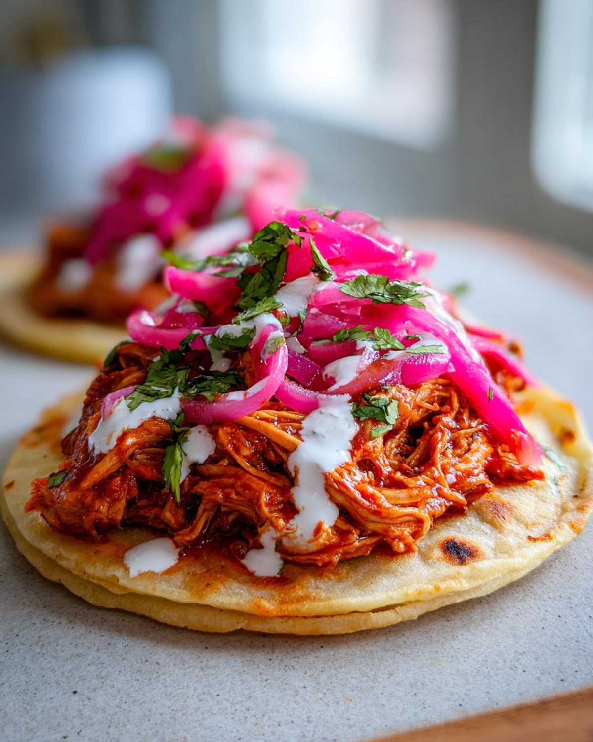 A close-up of a delicious Chicken Tinga Tostadas topped with vibrant pink pickled onions, crema, and cilantro.