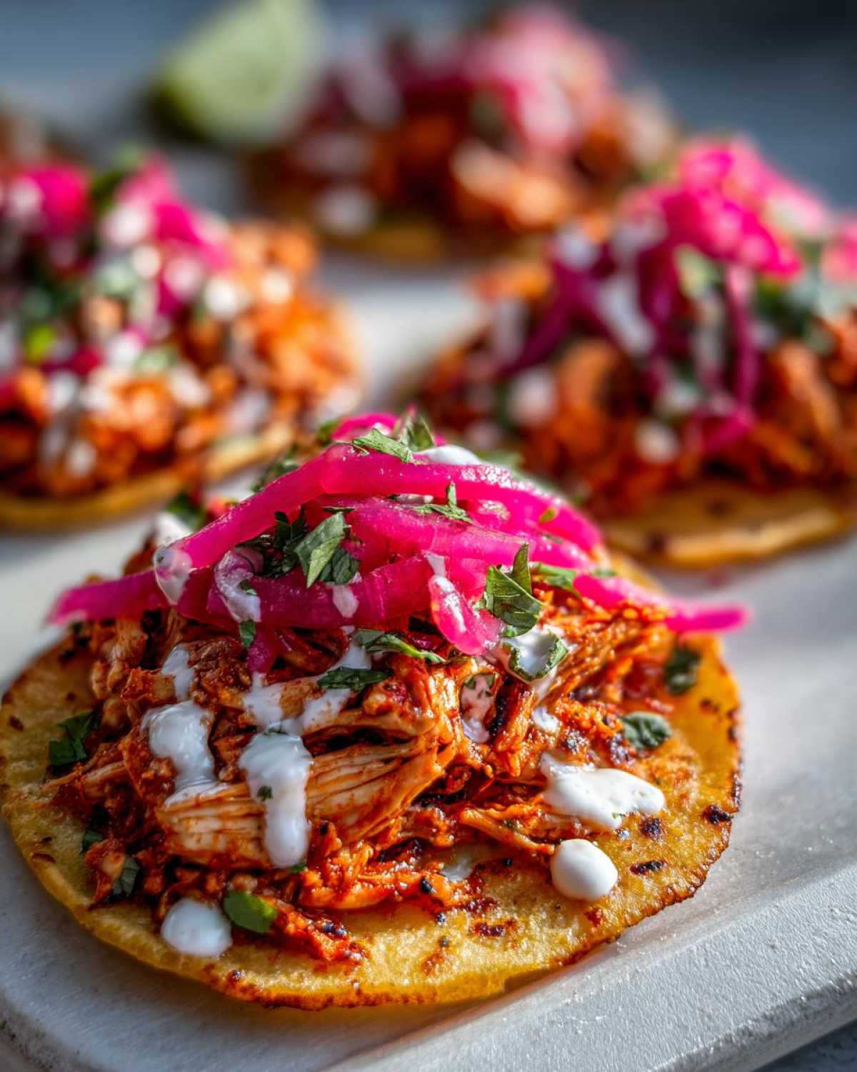 A close-up of a crispy tostada topped with shredded Chicken Tinga, drizzled with crema, and finished with bright pink pickled onions.